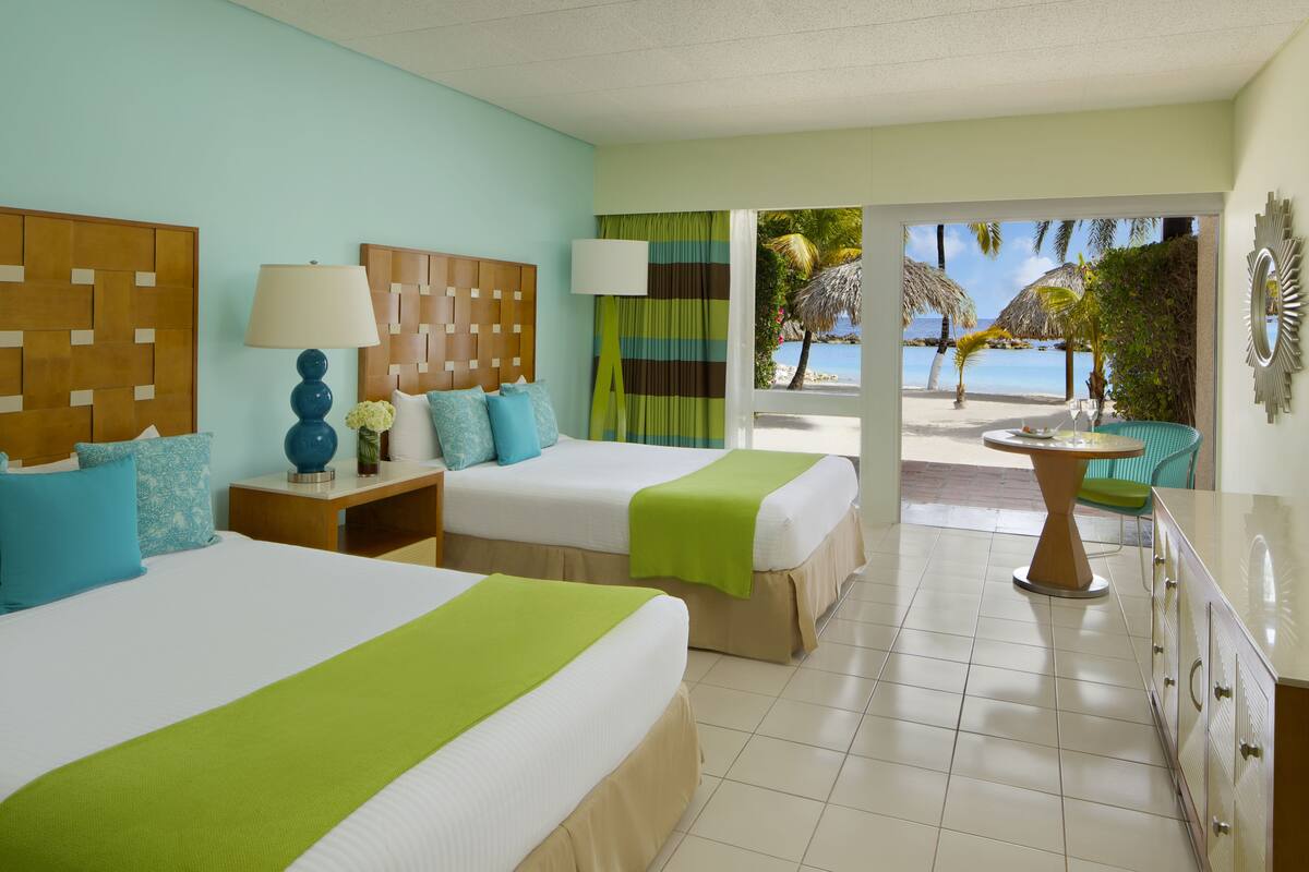 A hotel room with two beds, a nightstand, a lamp, a table, and a view of a beach.