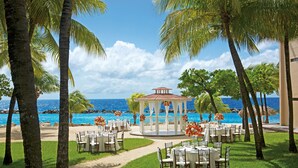 A gazebo with tables and chairs set up for an outdoor event, overlooking a beach with palm trees and the ocean.