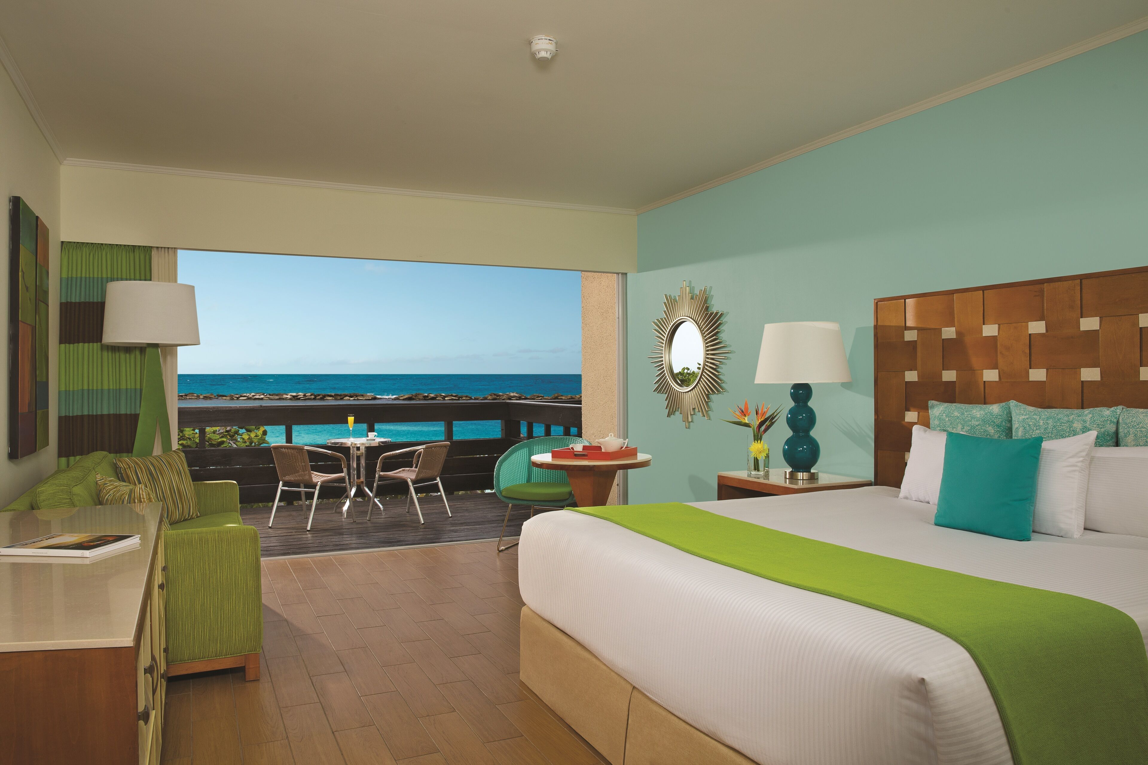 Ein Hotelzimmer mit einem großen Bett, einem grünen Sessel, einem kleinen Tisch und Blick aufs Meer.