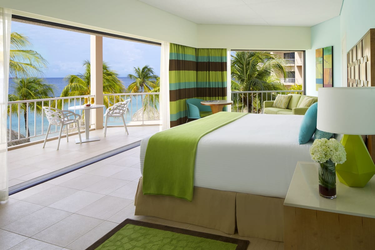 A hotel room with a balcony, a bed with a green bedspread, a small table with a vase of flowers, and a view of the ocean.