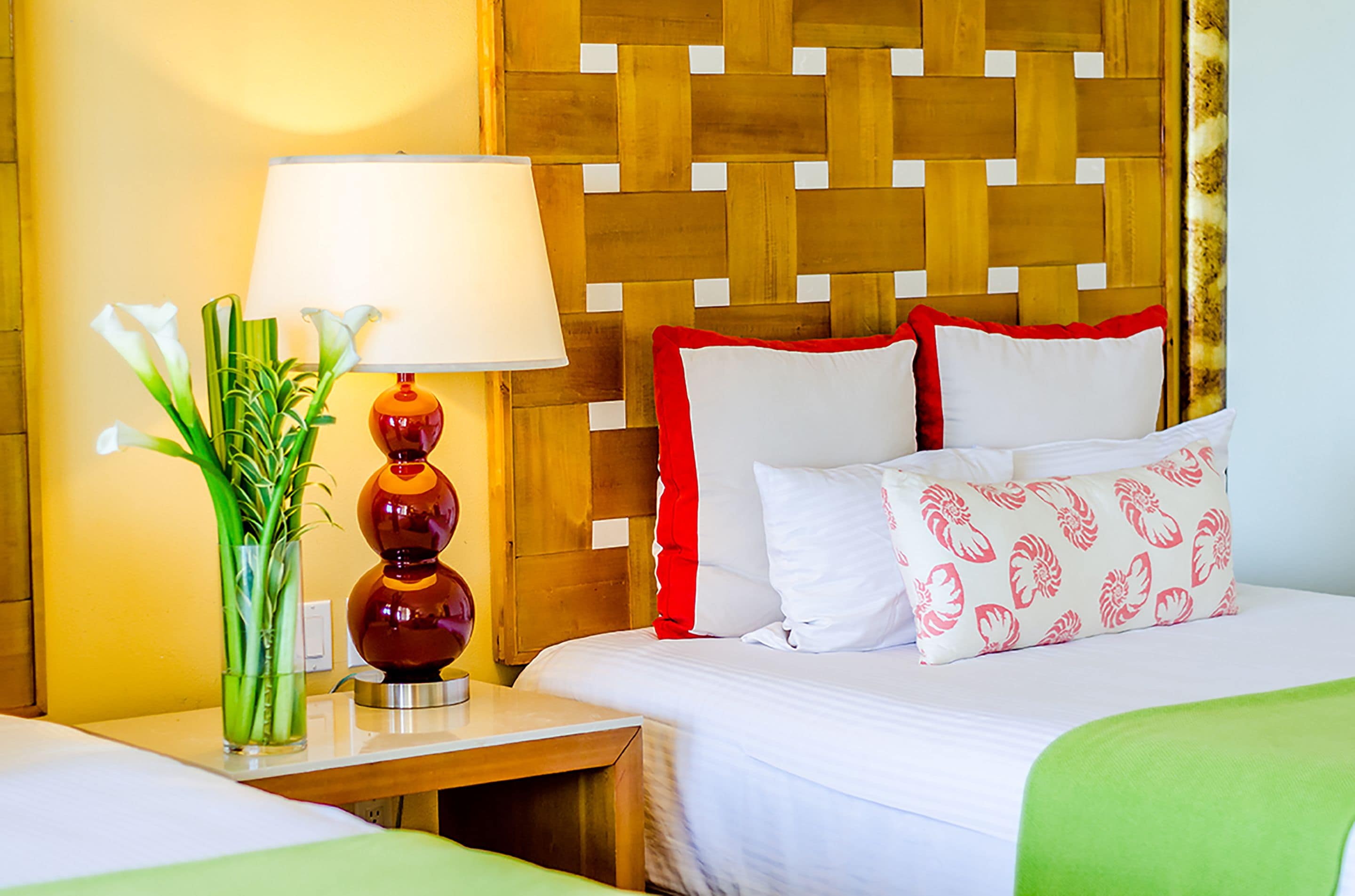 Habitación de hotel con cabecera de madera, una mesita de noche con lámpara, un jarrón con flores y una cama con ropa de cama blanca y roja.