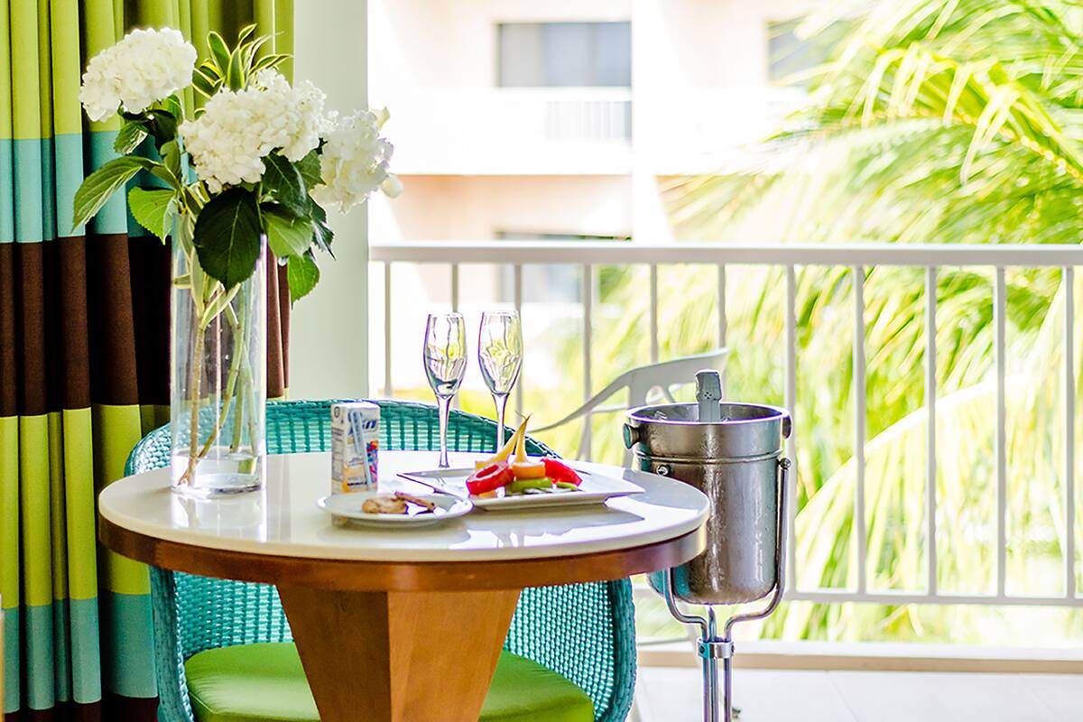 A dining table with a vase of flowers, two wine glasses, a plate of food, and a metal container, set on a balcony with a view of palm trees.