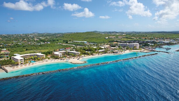 An aerial view of a coastal resort with multiple buildings, a sandy beach, and clear turquoise waters.