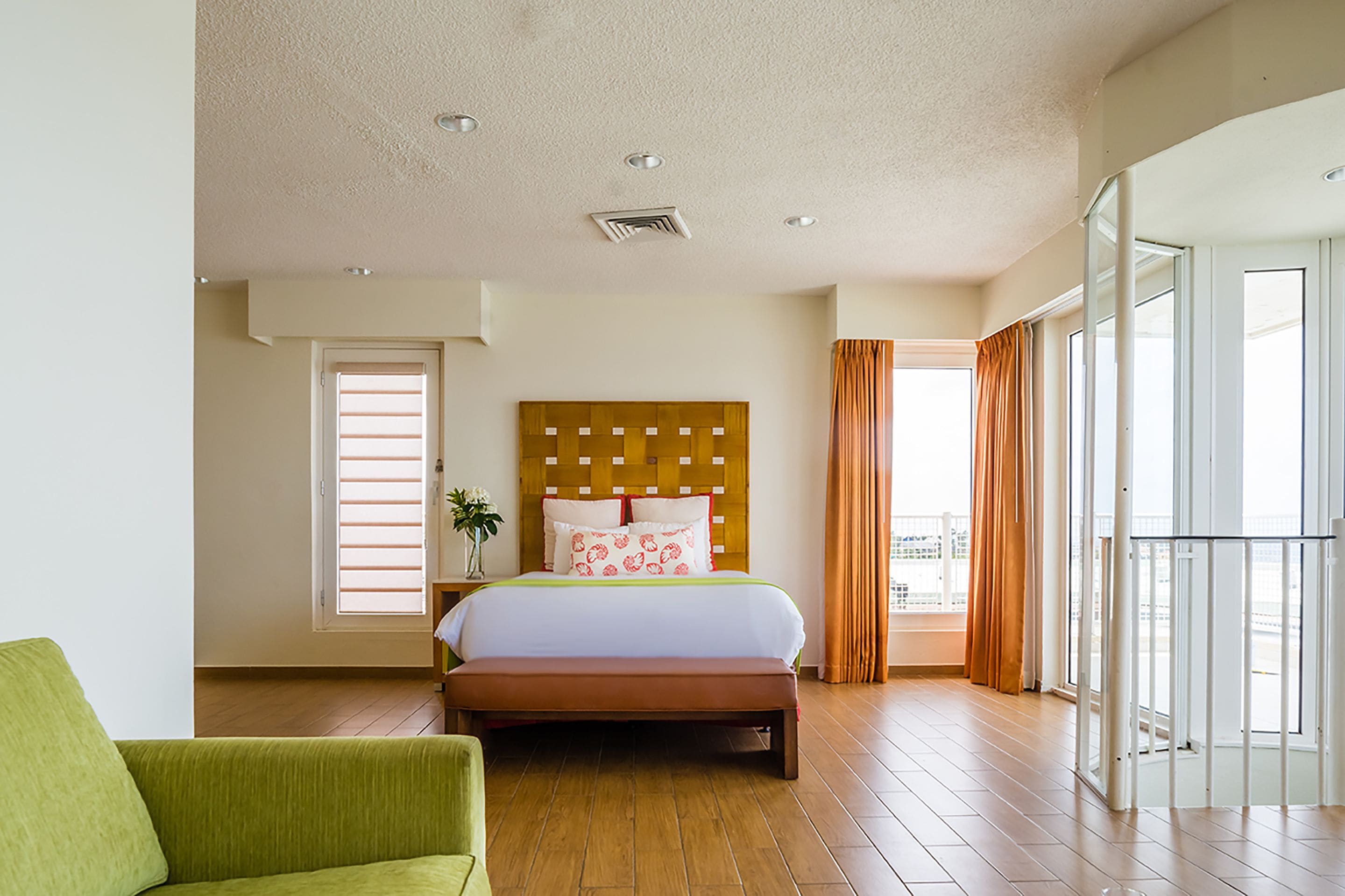 A hotel room with a wooden floor, a bed with a headboard, a green armchair, and a balcony with curtains.