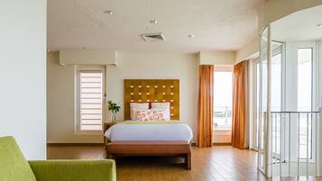 A hotel room with a wooden floor, a bed with a headboard, a green armchair, and a balcony with curtains.