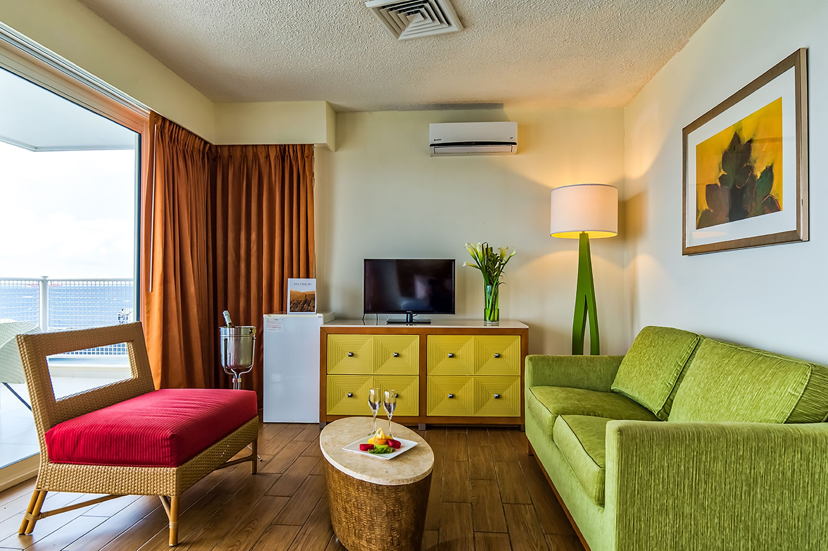 Habitación de hotel con balcón, televisión, sofá verde, silla de mimbre y una pequeña mesa redonda con una fuente de fruta y dos vasos.