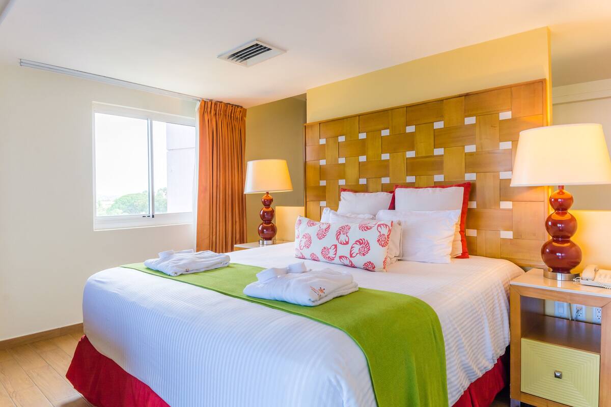 A hotel room with a large bed, two bedside lamps, a nightstand, a window with curtains, and a view of greenery outside.