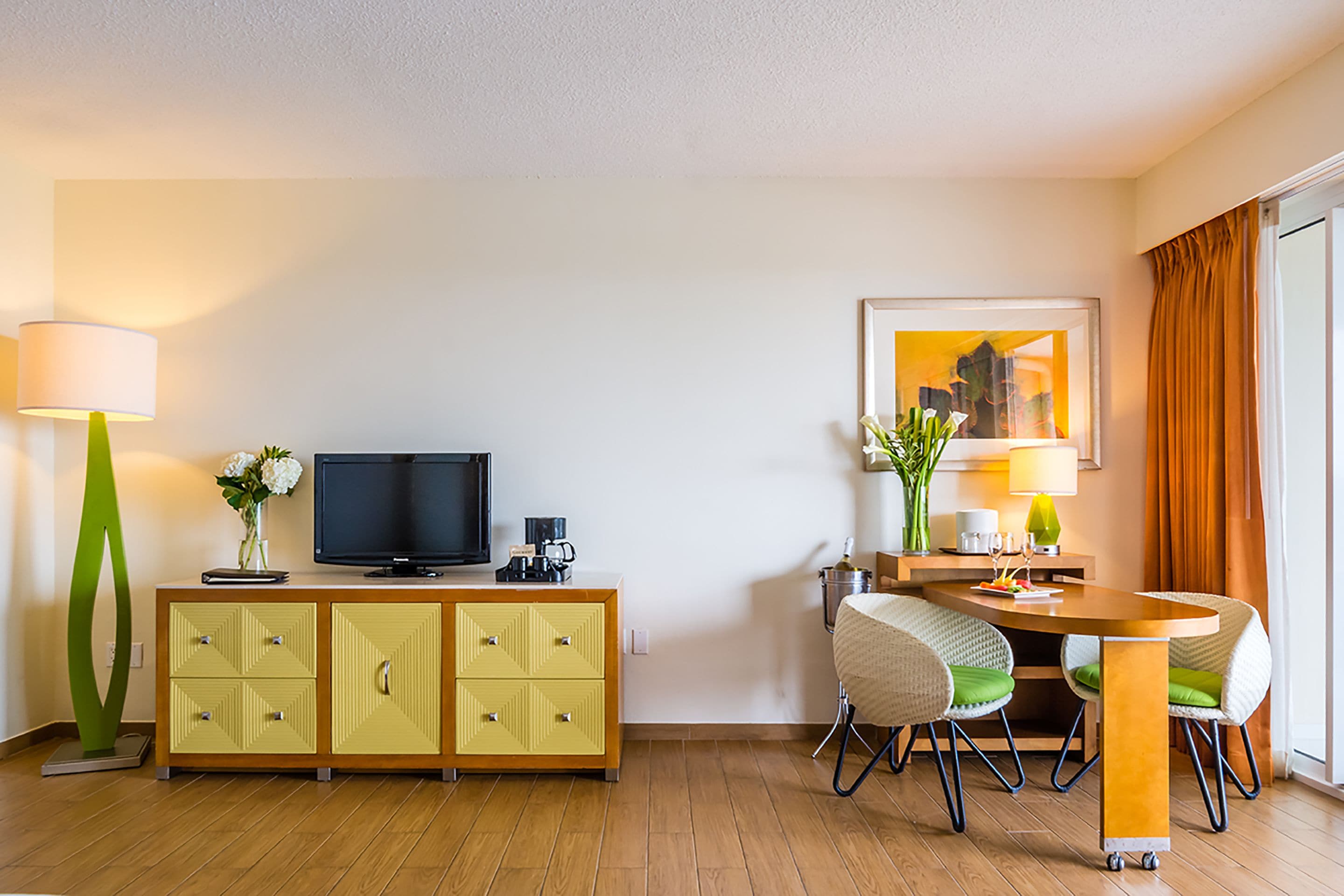 Een hotelkamer met een houten vloer, een geel kastje met lades, een flatscreen televisie, een eettafel met stoelen, een lamp en een ingelijste foto aan de muur.