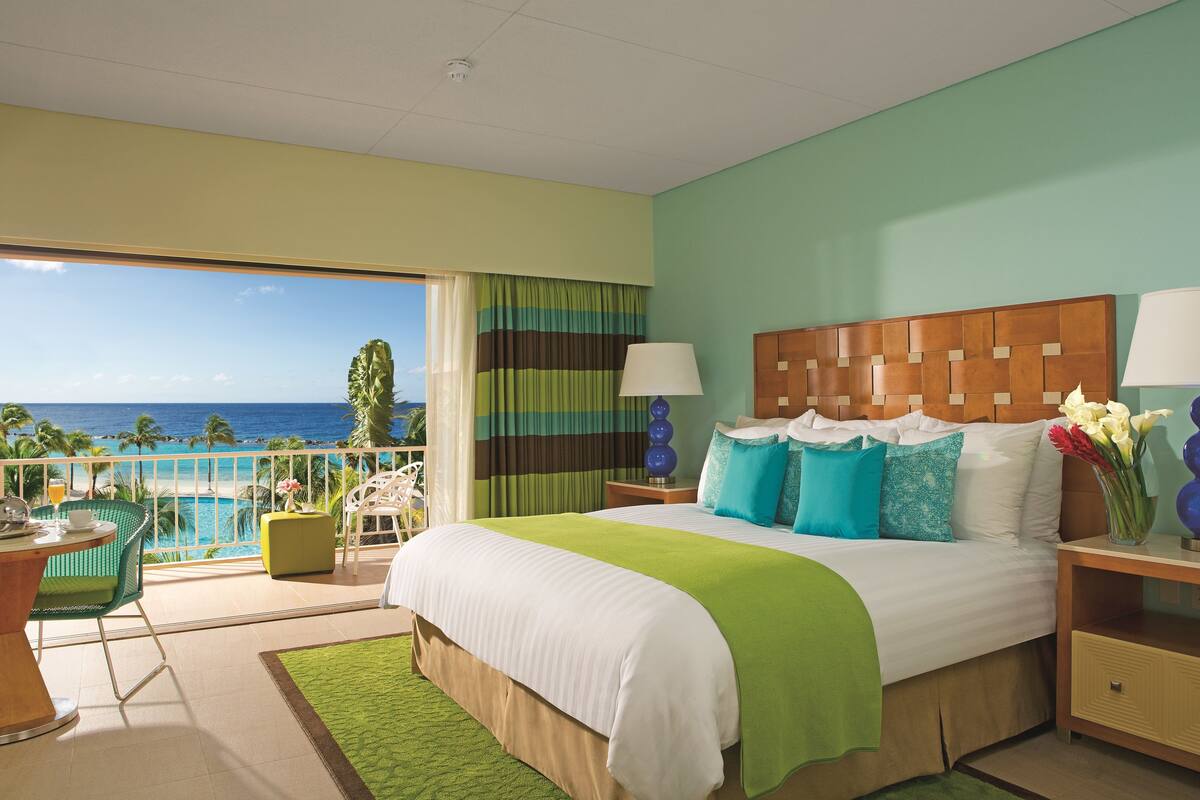 A hotel room with a large bed, a balcony with a view of the ocean, and a dining area with a table and chairs.
