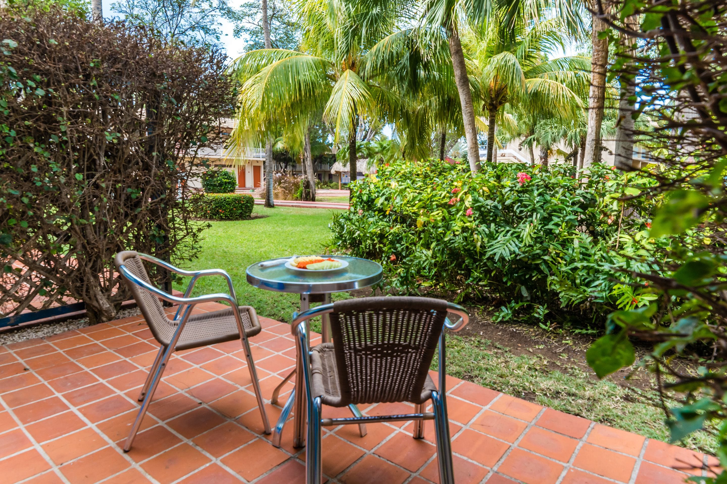 An outdoor patio with two metal chairs and a small round table, surrounded by lush greenery and palm trees.