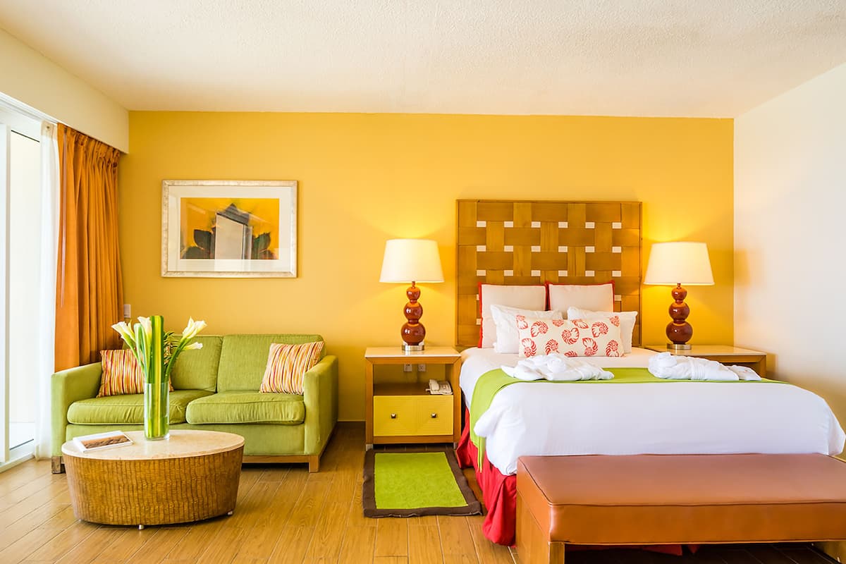 A hotel room with a green sofa, a round coffee table, a bed with white bedding, two bedside lamps, and a framed picture on the wall.