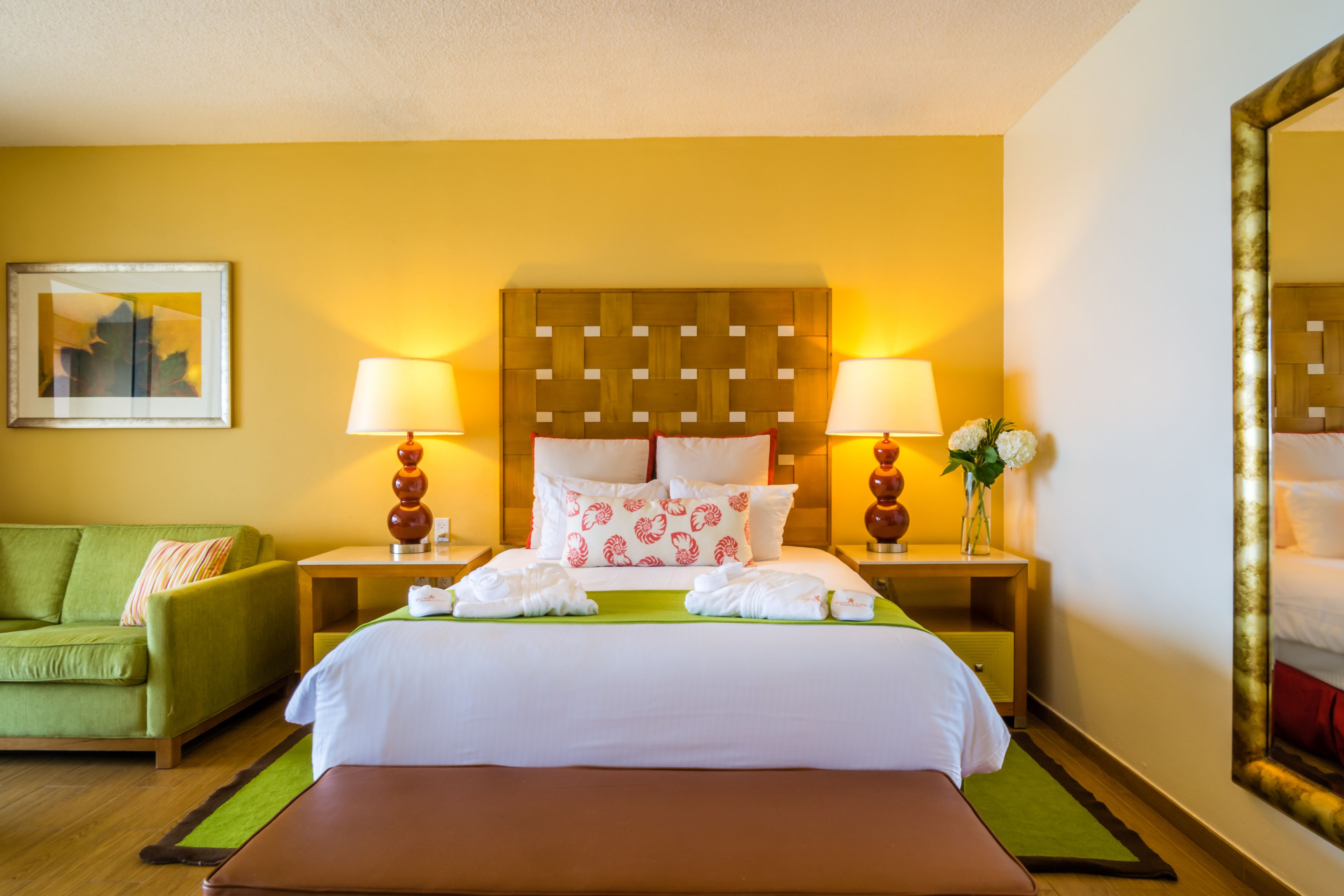 A hotel room with a bed, two bedside lamps, a green sofa, a small table, and a mirror.