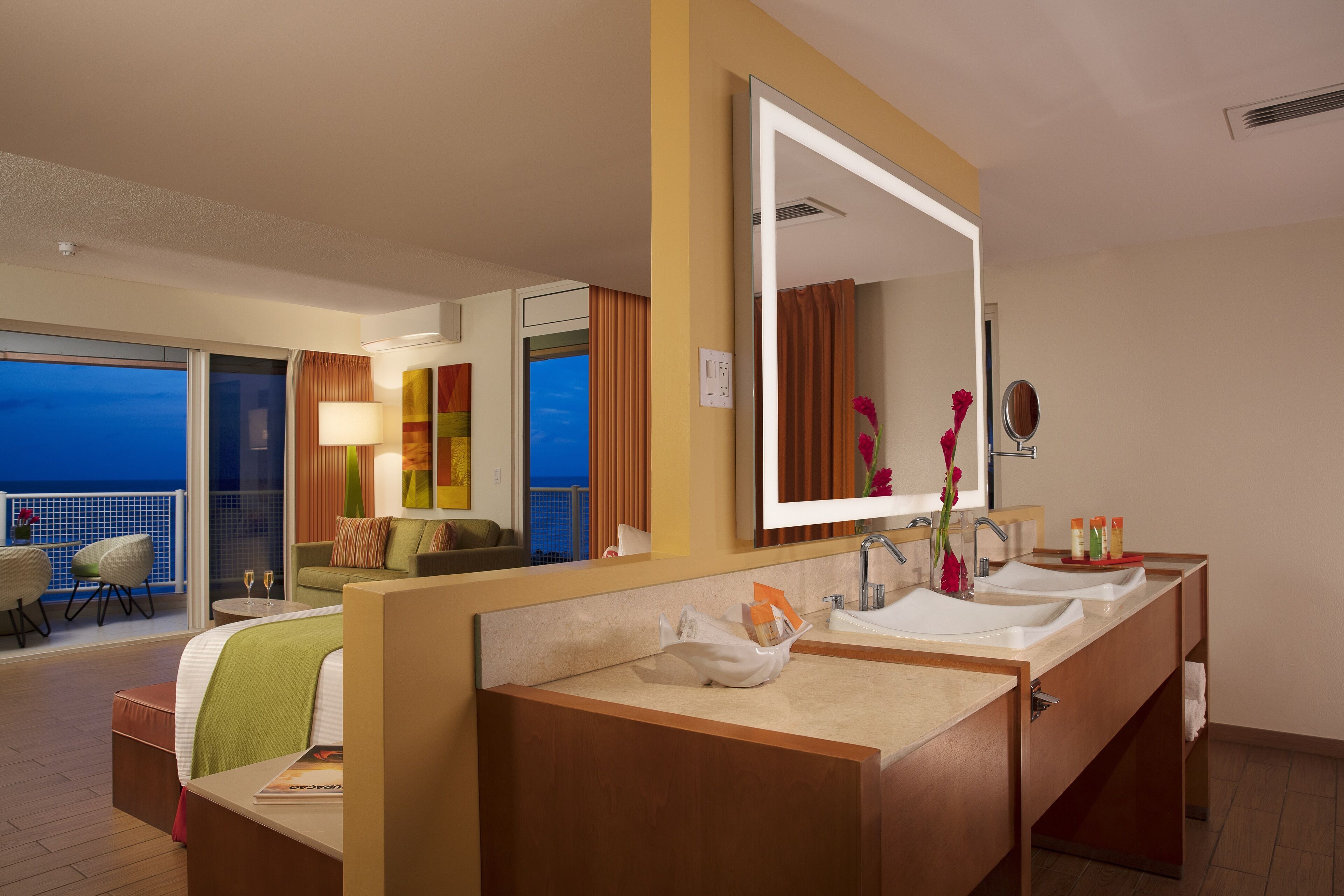 A hotel room with a balcony, a large mirror, a double sink vanity, and a view of the city at dusk.