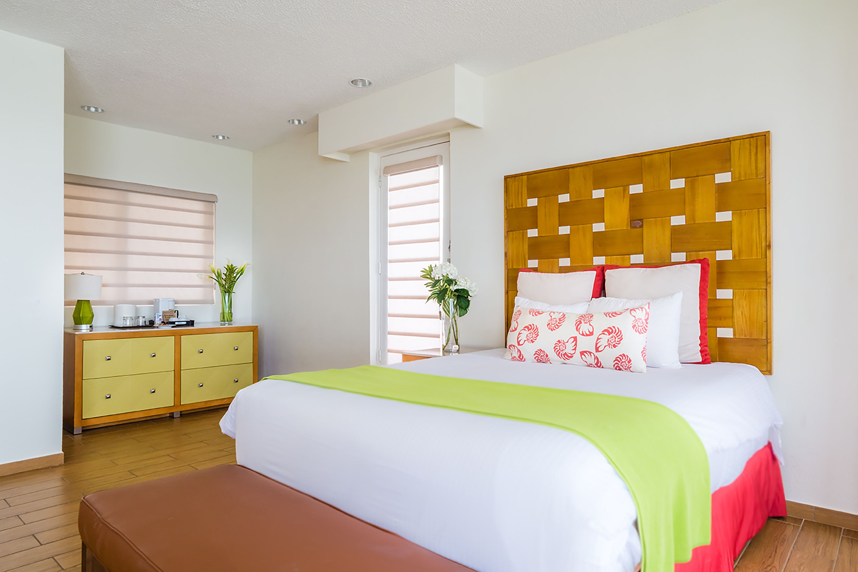 Hotelkamer met een bed, houten hoofdbord, nachtkastje, dressoir en raam met jaloezieën.
