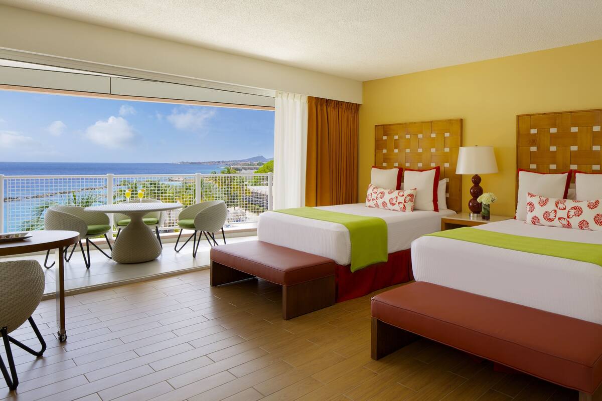 A hotel room with two beds, a balcony with a view of the sea, a dining area with chairs, and a small table.