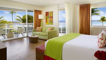 Habitación de hotel con cama, sofá verde, mesa de comedor con sillas y balcón con vista al océano y palmeras.
