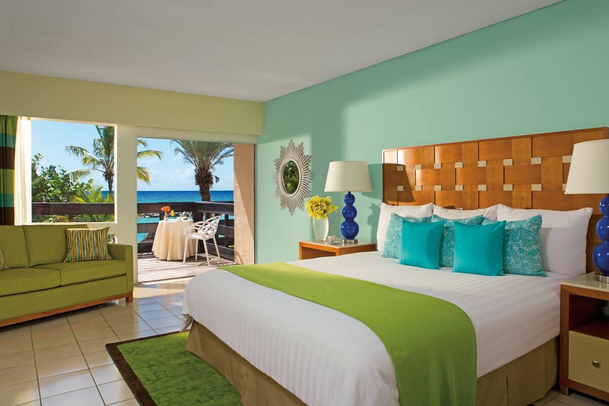 A hotel room with a bed, a green sofa, a balcony with a view of the ocean, and a table with chairs.
