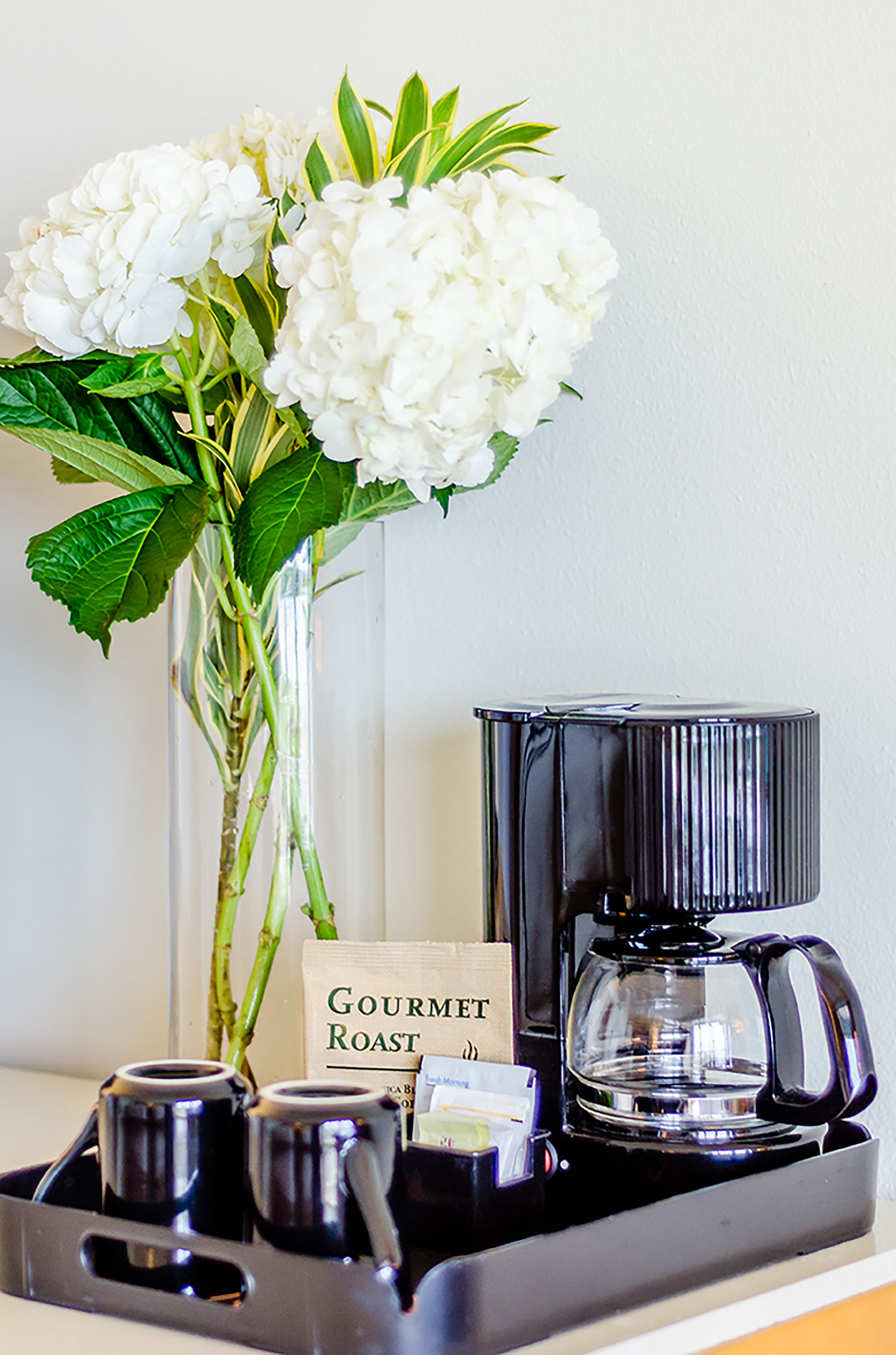 A coffee maker, a tray, and a vase with white flowers and green leaves.