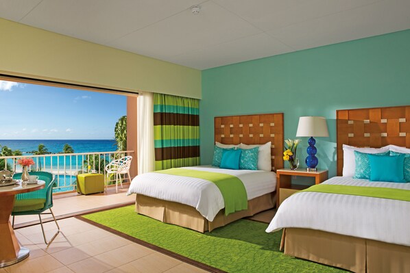 A hotel room with two beds, a balcony with a view of the ocean, and a small dining area.