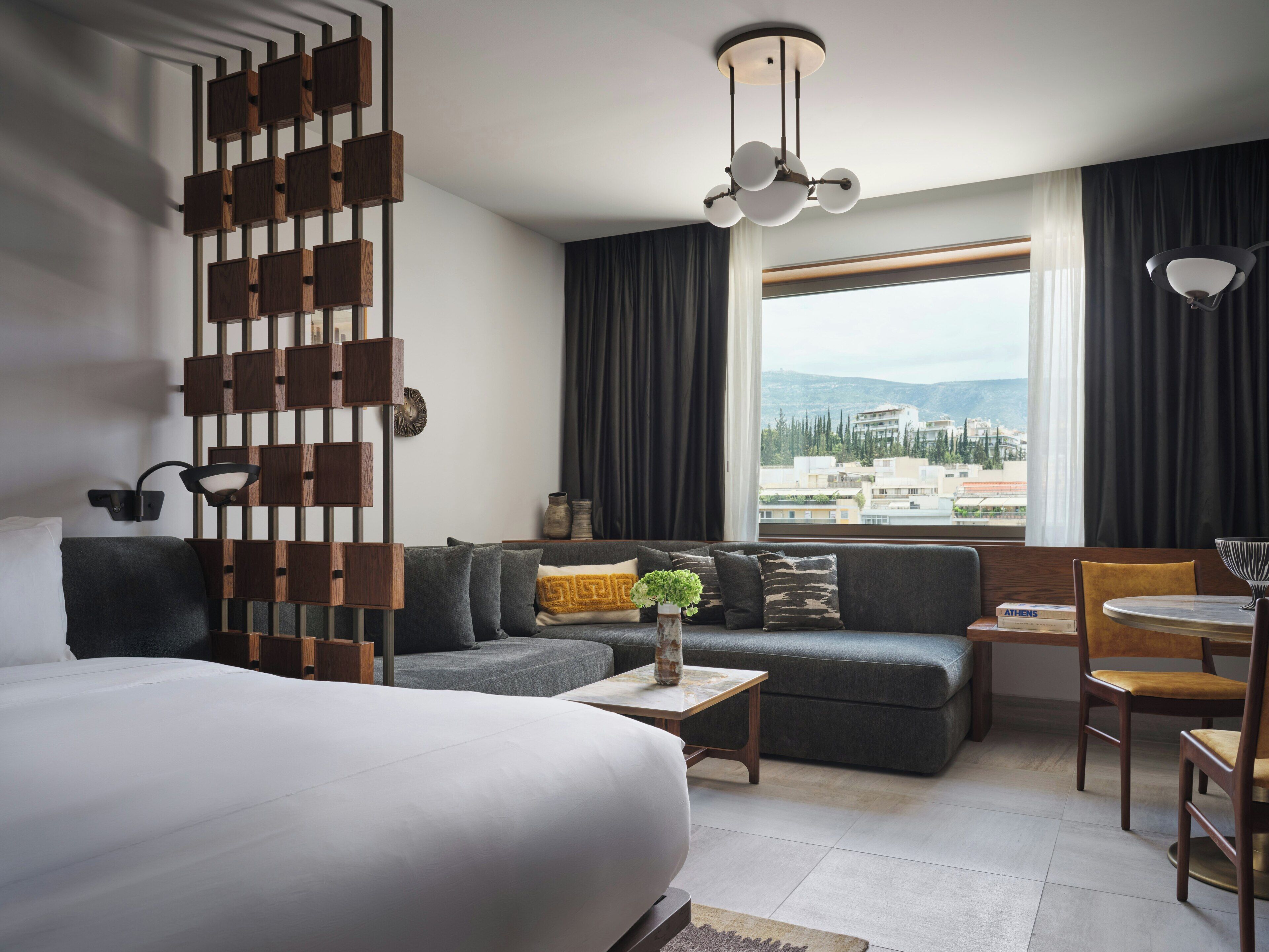 A modern hotel room with a large bed, a sofa, a dining area, and a view of the city.