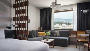 A modern hotel room with a large bed, a sofa, a dining area, and a view of the city.