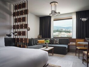 A modern hotel room with a large bed, a sofa, a dining area, and a view of the city. - Hilton Athens (Athens)