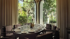 A dining area with a round table set for six, surrounded by upholstered chairs, large windows with a view of greenery, and a chandelier with multiple light fixtures. - Hilton Athens (Athens)