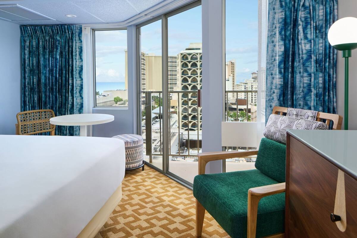 A hotel room with a bed, a green armchair, a small round table, and a view of the cityscape through large windows.