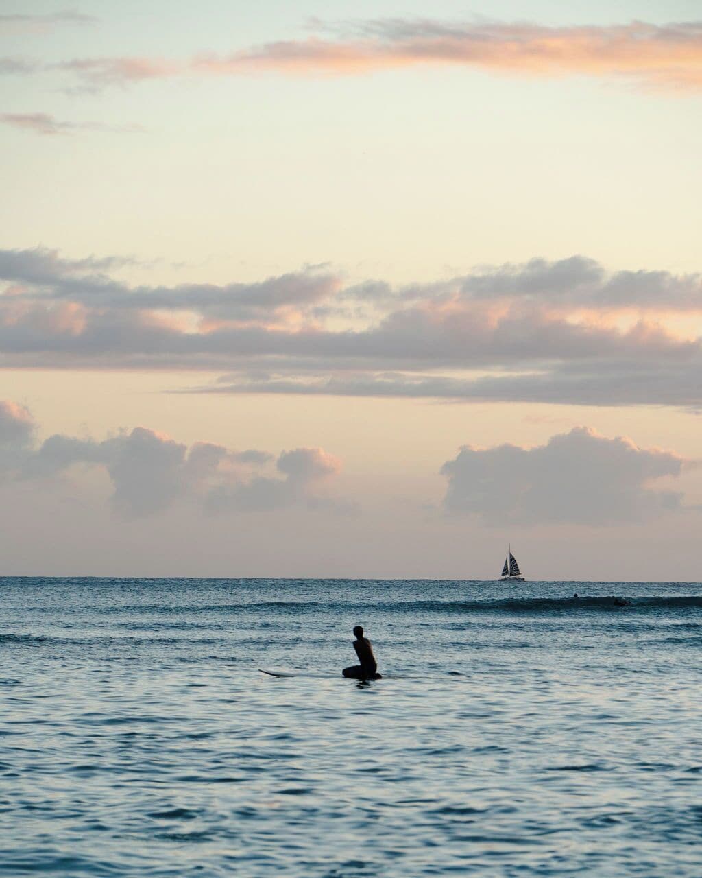 Mar en calma con un velero a lo lejos y una persona sentada en una tabla de surf al frente.