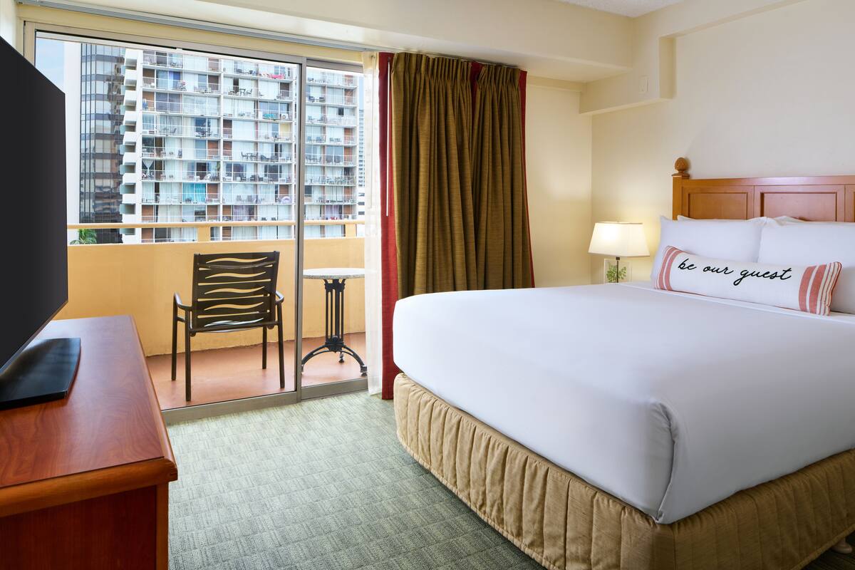 A hotel room with a large bed, a desk with a TV, a chair, a window with a view of a high-rise building, and a sign on the bed that says "be our guest".