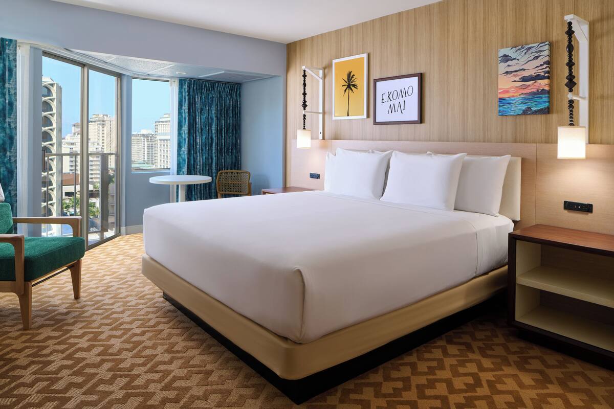 A hotel room with a large bed, a small table, a chair, and a view of the city through the window.