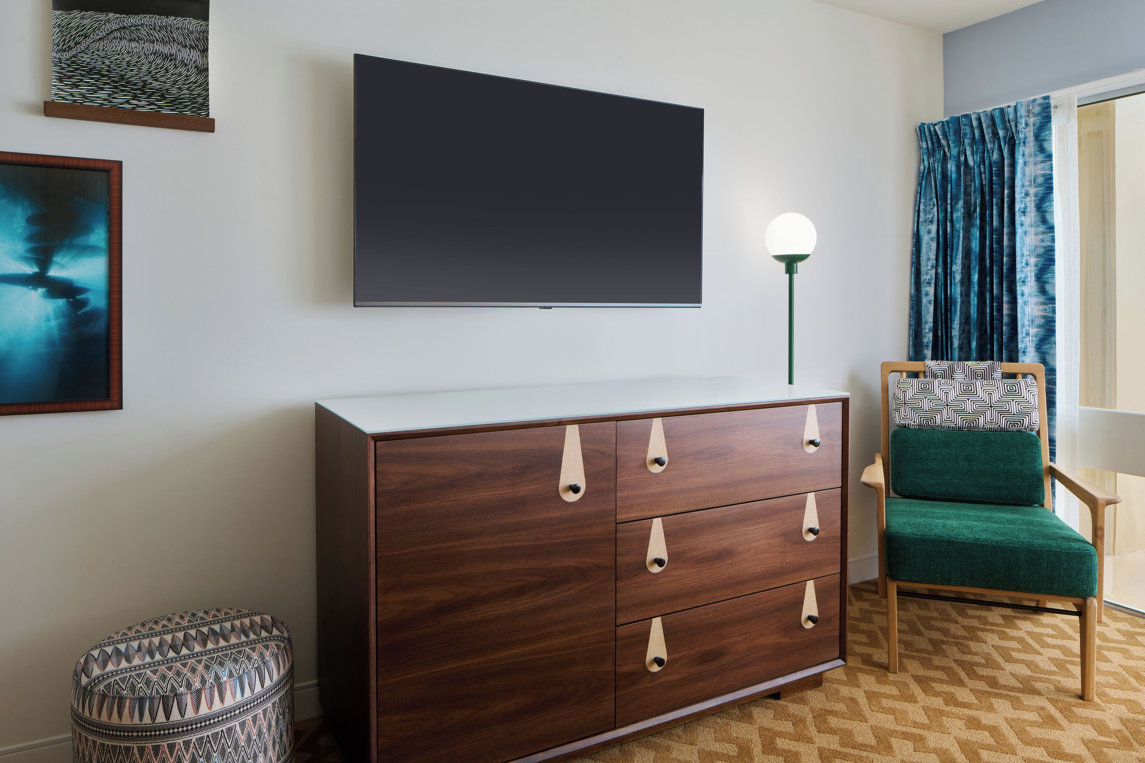 Una habitación moderna con un televisor de pantalla plana grande montado en la pared, un aparador de madera, un sillón tapizado de color verde y un reposapiés estampado.