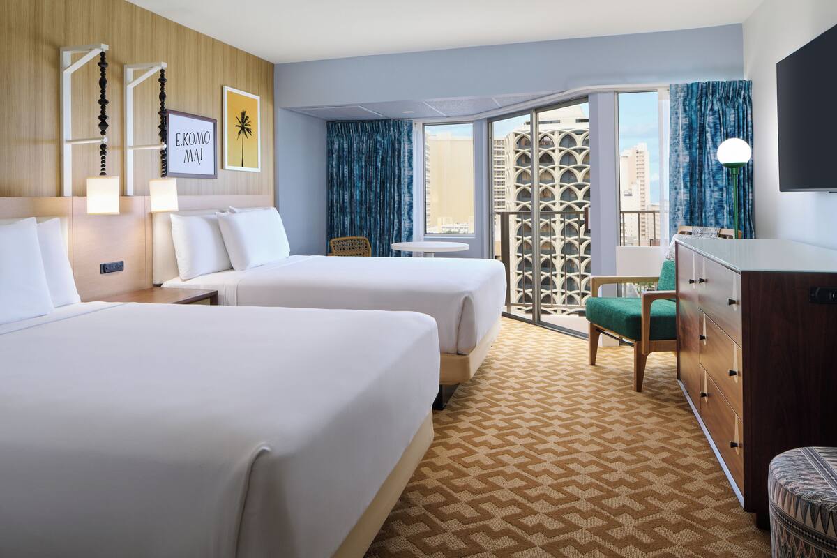 A hotel room with two beds, a wooden headboard, a small table, a chair, a dresser, and a balcony with a view of the city.