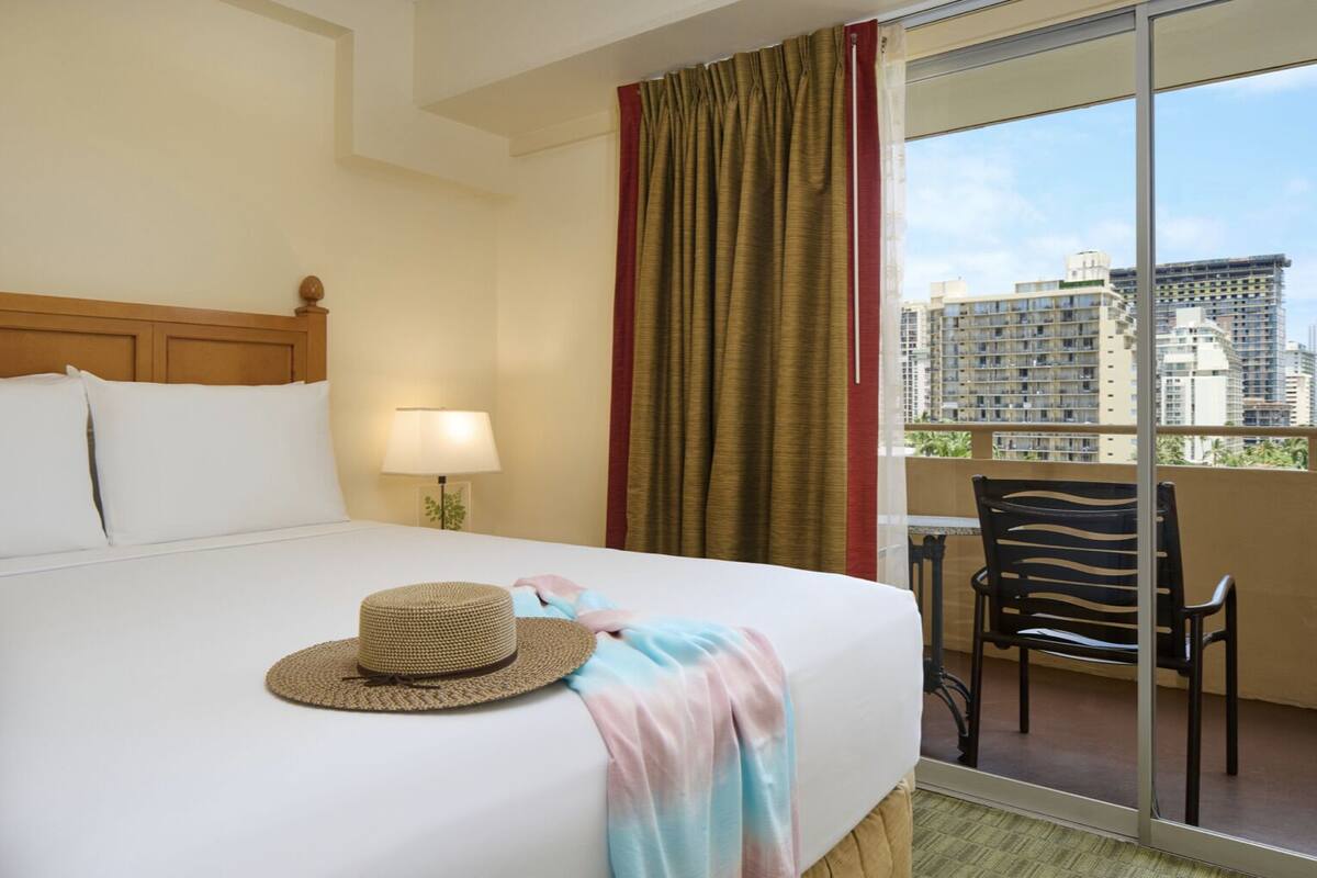 A hotel room with a bed, a lamp, a hat, a chair, and a balcony with a city view.