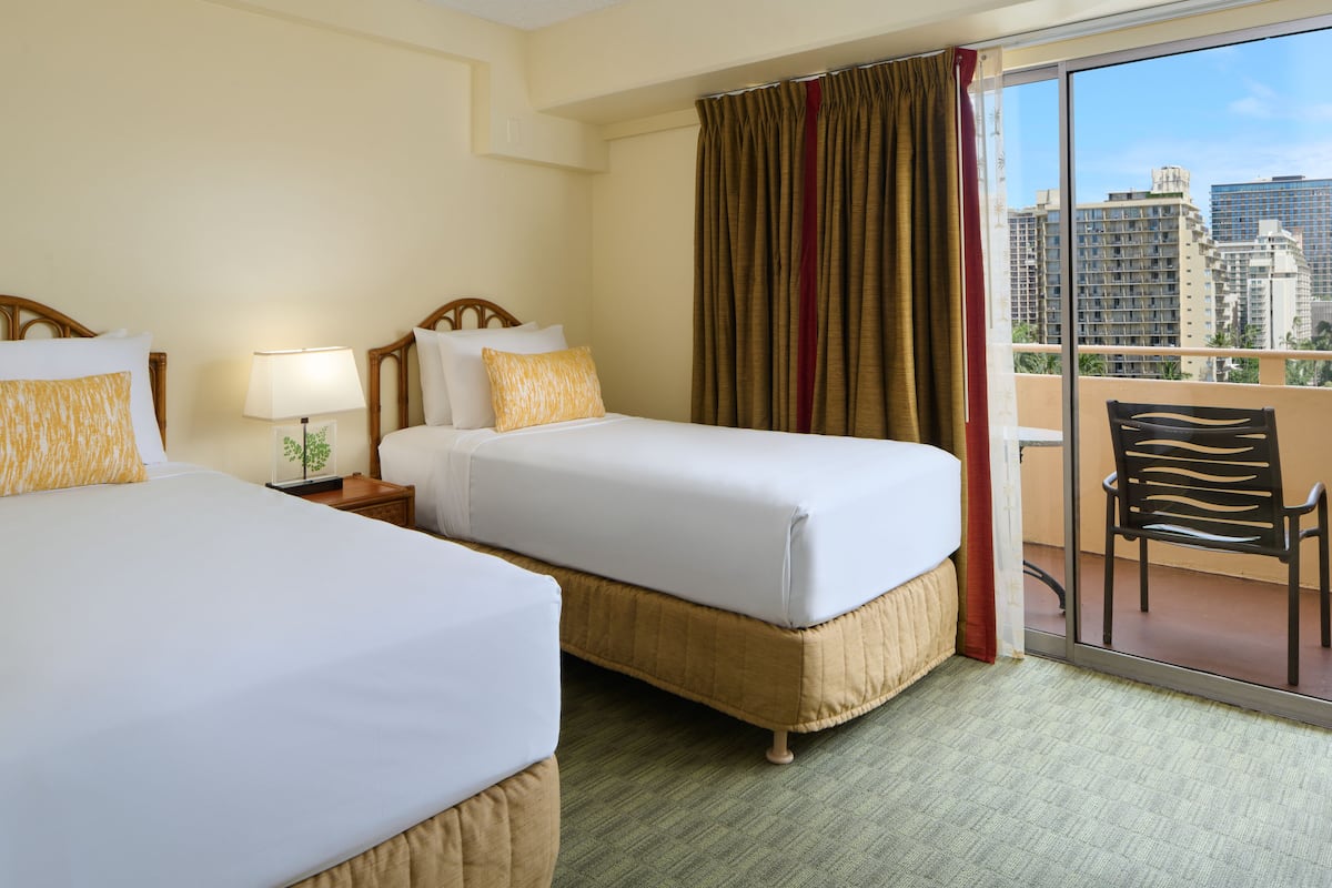 A hotel room with two beds, a balcony with a chair, and a view of city buildings.