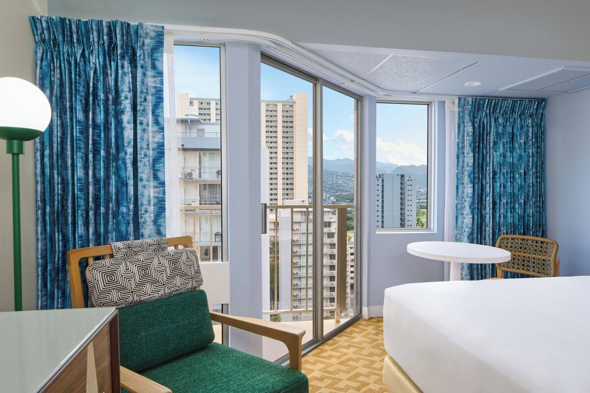 A hotel room with a bed, a chair, a small table, and a view of tall buildings through large windows.