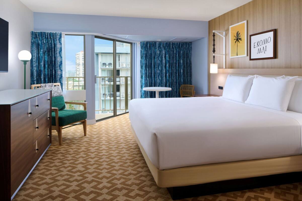 A hotel room with a large bed, a desk, a chair, a small table, and a view of the outside.