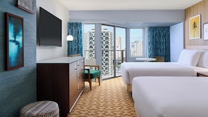 Ein Hotelzimmer mit einem großen Bett, einem Schreibtisch, einem Sessel, einem kleinen Hocker, einem Fernseher und einem Balkon mit Blick auf die Stadt.