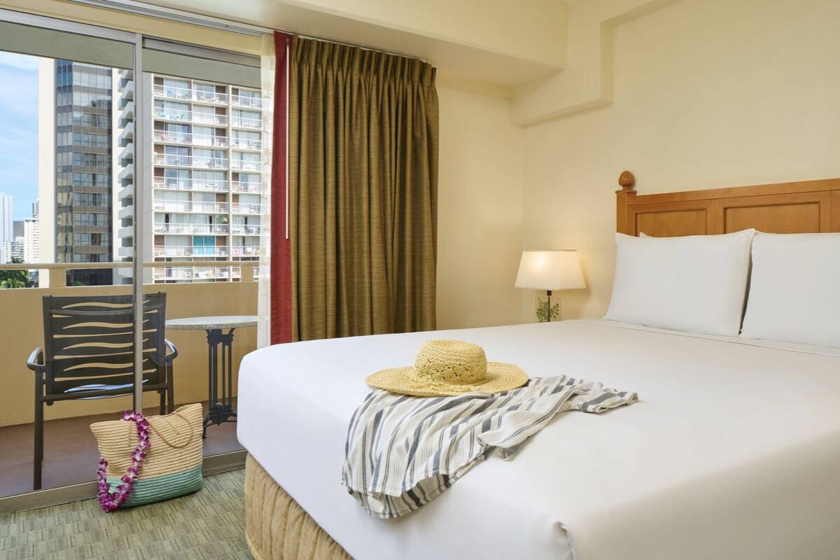 A hotel room with a bed, a chair, a table, a lamp, and a view of buildings.