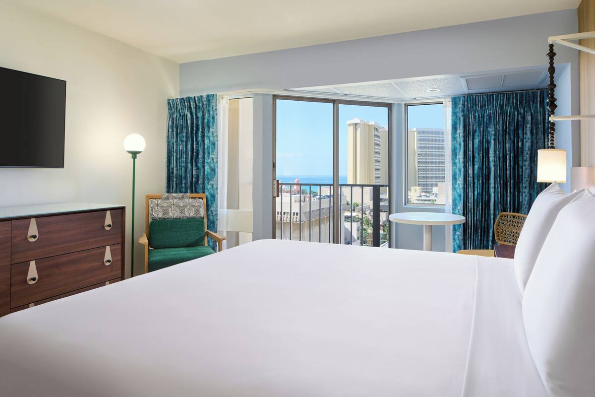 A hotel room with a large bed, a desk, a chair, a TV, and a balcony with a view of the city.