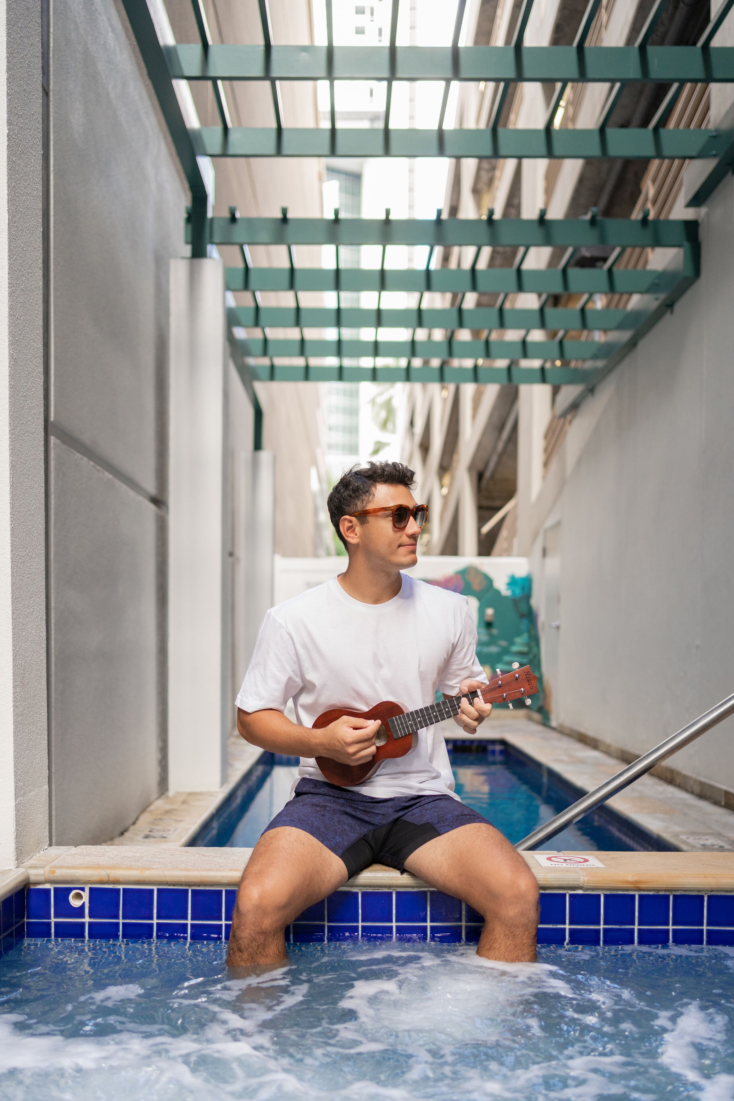 Una persona sentada en un pequeño área de piscina con barandilla de metal, sosteniendo un ukelele.
