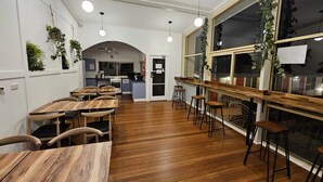 A restaurant with wooden tables and chairs, a kitchen in the background, and hanging plants. - Waterview Gosford Motor Inn (West Gosford)