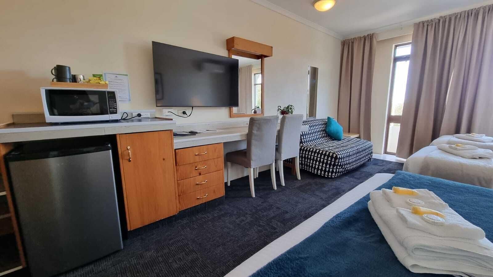 Une chambre d’hôtel équipée d’une kitchenette, d’un bureau avec un téléviseur à écran plat, d’une chaise, d’un lit avec des serviettes et d’une fenêtre avec des rideaux.