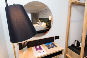 Ett hotellrum med en säng, en rund spegel, ett skrivbord med broschyrer och en kaffebryggare.