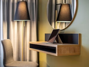 Ein modernes Hotelzimmer mit einem Nachttisch aus Holz, einem Laptop, einer Lampe, einem Sessel und einem Spiegel an der Wand.