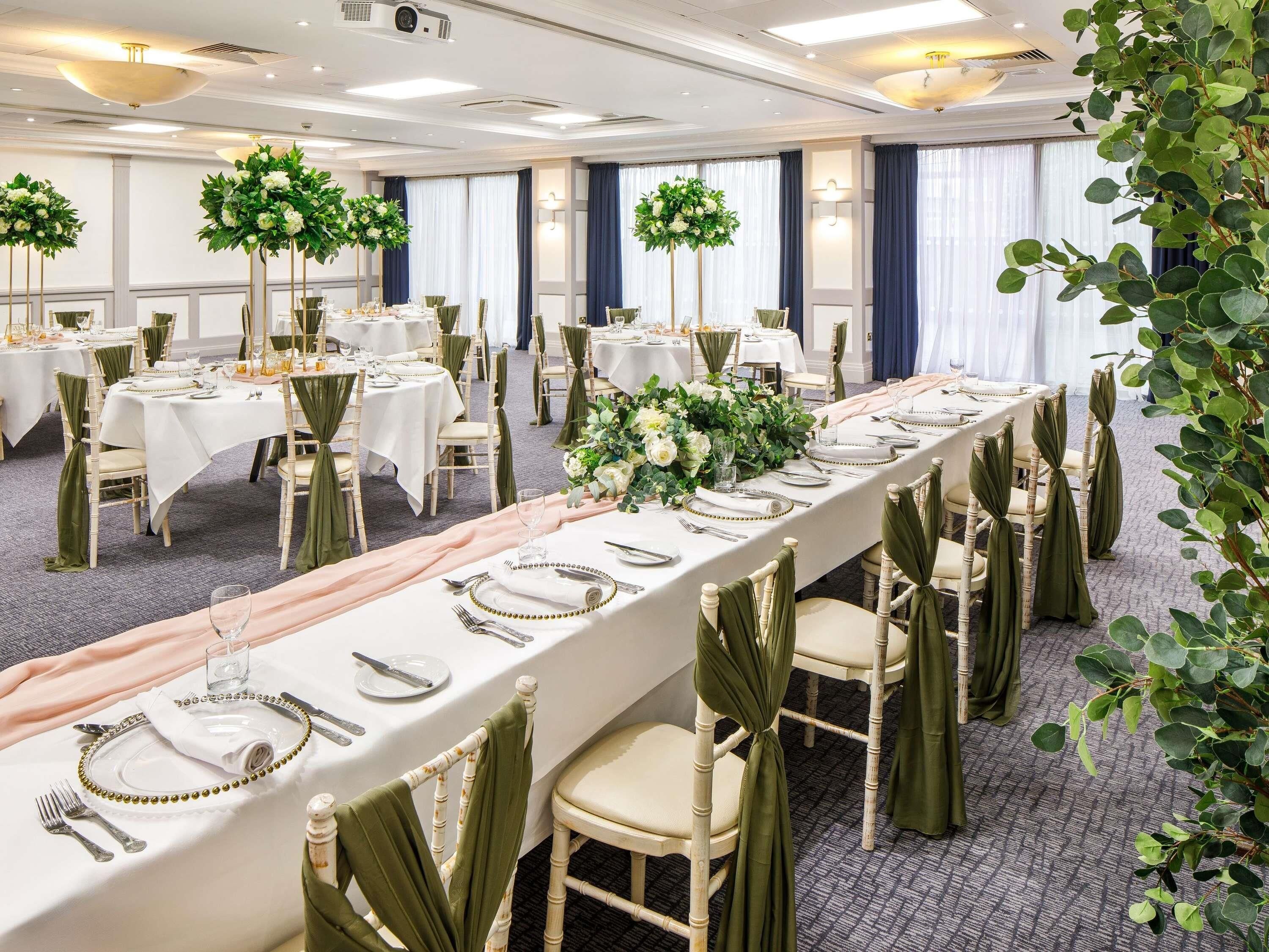 A banquet hall with long tables set for dining, surrounded by chairs with green sashes, and decorated with floral centerpieces.