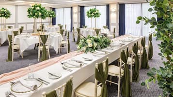 A banquet hall with long tables set for dining, surrounded by chairs with green sashes, and decorated with floral centerpieces.