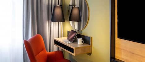 A hotel room with a bed, a desk with a lamp, a chair, a mirror, and a TV.
