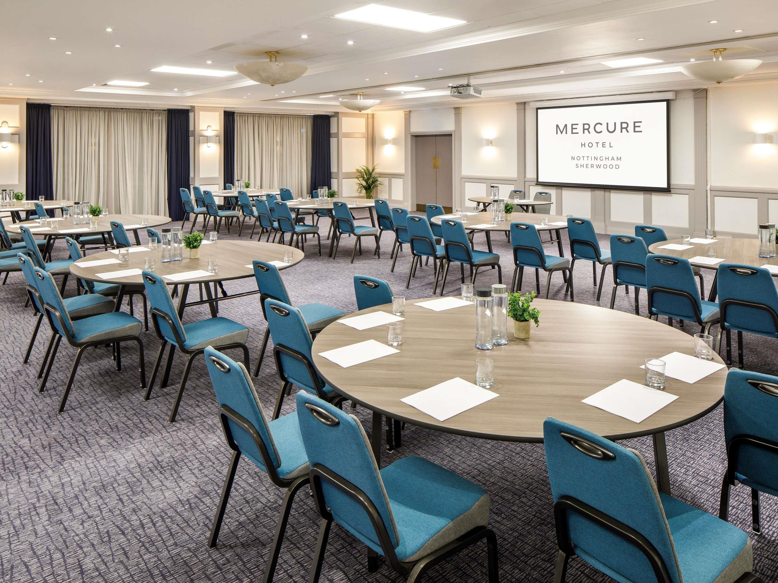 A conference room with round tables and blue chairs, a projector screen displaying 'Mercure Hotel Nottingham Sherwood', and a carpeted floor.