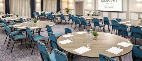 A conference room with round tables and blue chairs, a projector screen displaying 'Mercure Hotel Nottingham Sherwood', and a carpeted floor.